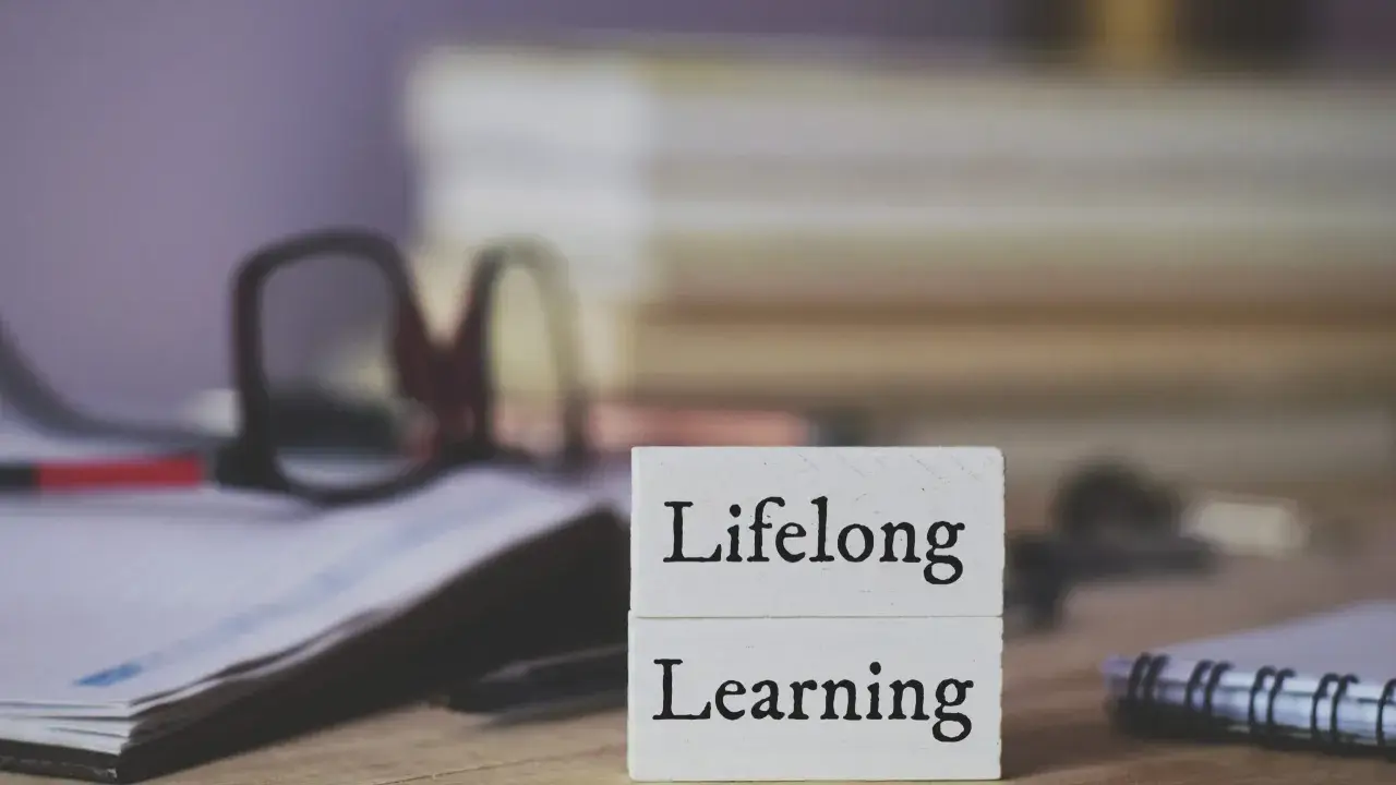 Dwa białe klocki z napisami "Lifelong" i "Learning" symbolizują uczenie się przez całe życie. W tle okulary i notatnik.
