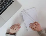 Ręce piszą notatki do CV po niemiecku przy laptopie, obok filiżanka kawy.