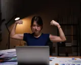 Młoda kobieta w godzinach nocnych 2026, siedząc przy biurku, pokazuje bicepsy.