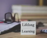 Dwa białe klocki z napisami "Lifelong" i "Learning" symbolizują uczenie się przez całe życie. W tle okulary i notatnik.