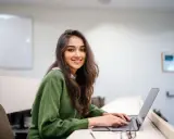 Uśmiechnięta studentka pracuje na laptopie w sali wykładowej. Jej status ucznia jest widoczny w otoczeniu i skupieniu na nauce.