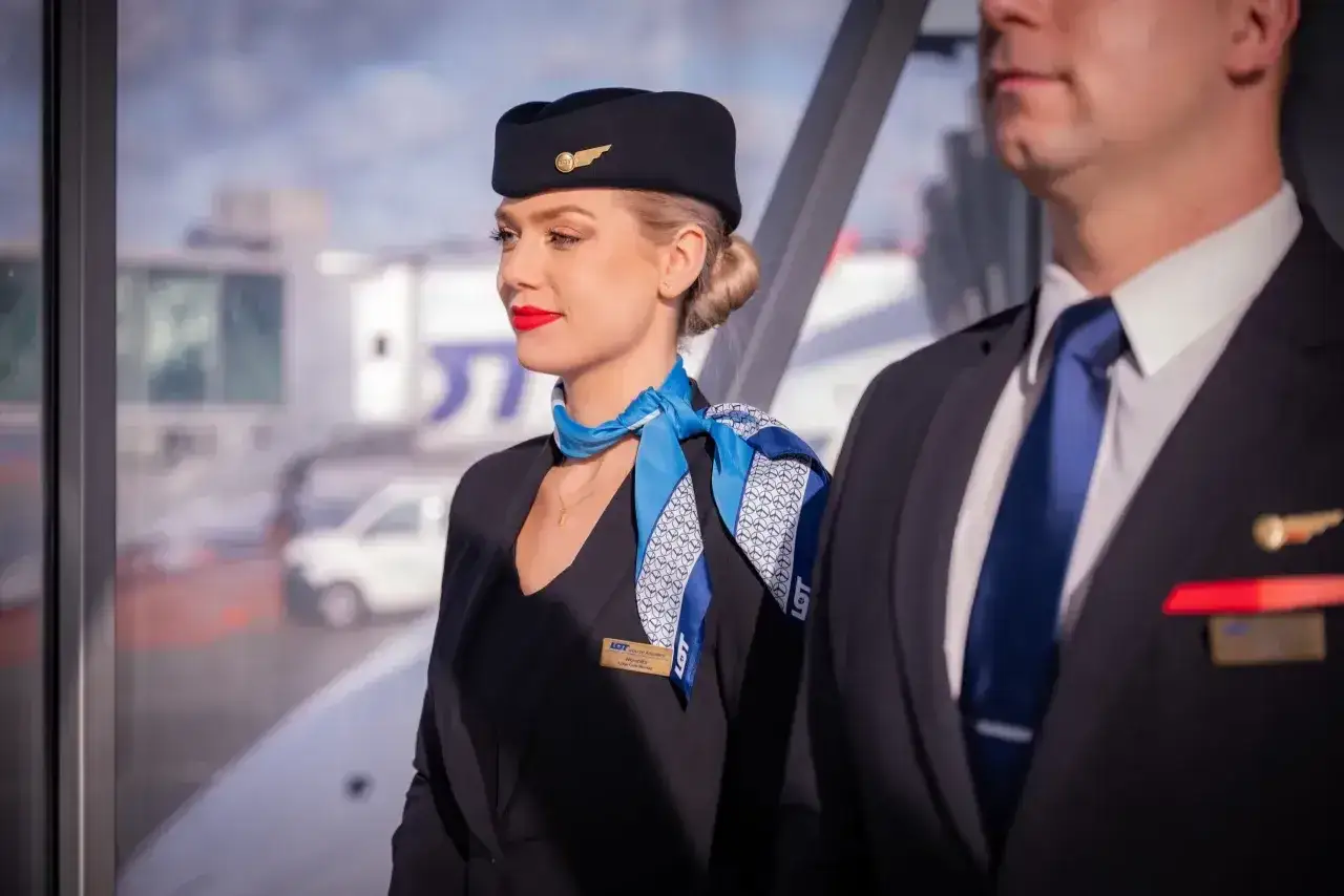 Uśmiechnięta stewardessa w granatowym mundurze i niebieskim z białym wzorem z logo LOT, gotowa do pracy. Obok niej stoi mężczyzna w garniturze.